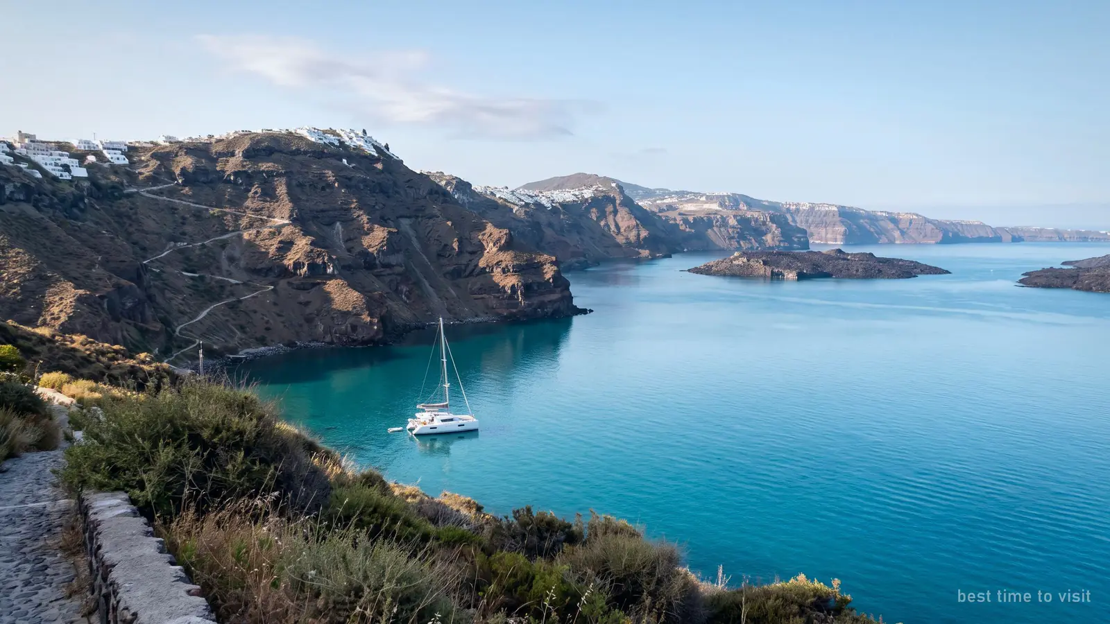 A catamaran at anchor in calm turquoise water below the Santorini caldera cliffs on a summer morning