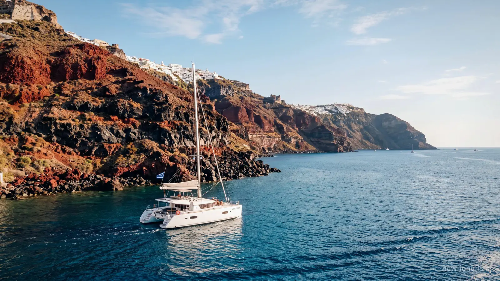 A catamaran sailing past the red and black volcanic cliffs of southern Santorini