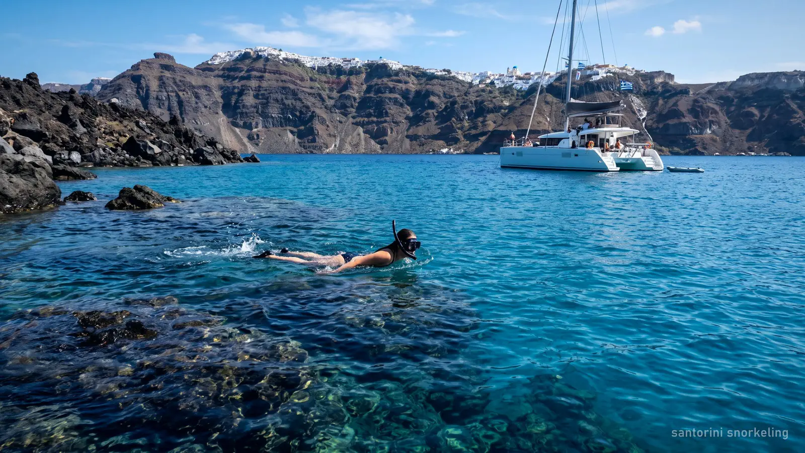 A snorkeller over dark volcanic rocks in clear water off Santorini, a catamaran anchored nearby